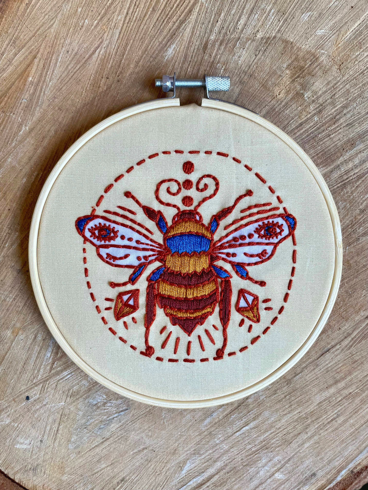 The Tangled Up In Hue 5 Beginner Winking Bee Embroidery Kit features a colorful bee with detailed wings and decorative patterns—ideal for beginner crafters or as display art on a wooden surface.