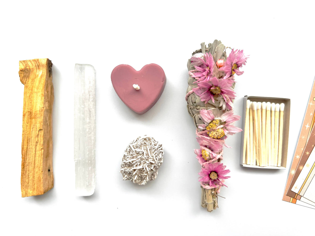 A flat lay on white displays The 12th Moon’s Goddess Intentions Self-Care Ritual Kit, featuring a Prairie Sage Bundle with pink flowers, Palo Santo stick, selenite wand, pink heart candle, crystal cluster, matches, and art prints.