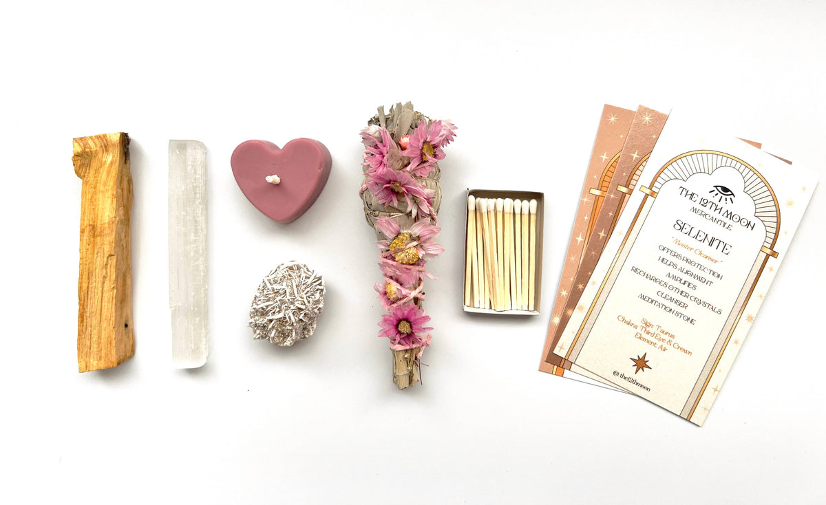 The 12th Moon’s Goddess Intentions Self-Care Ritual Kit features a Palo Santo stick, selenite wand, heart-shaped pink candle, Prairie Sage Bundle, white crystal, matches, and info cards—all artfully arranged to channel Divine Feminine Energy.