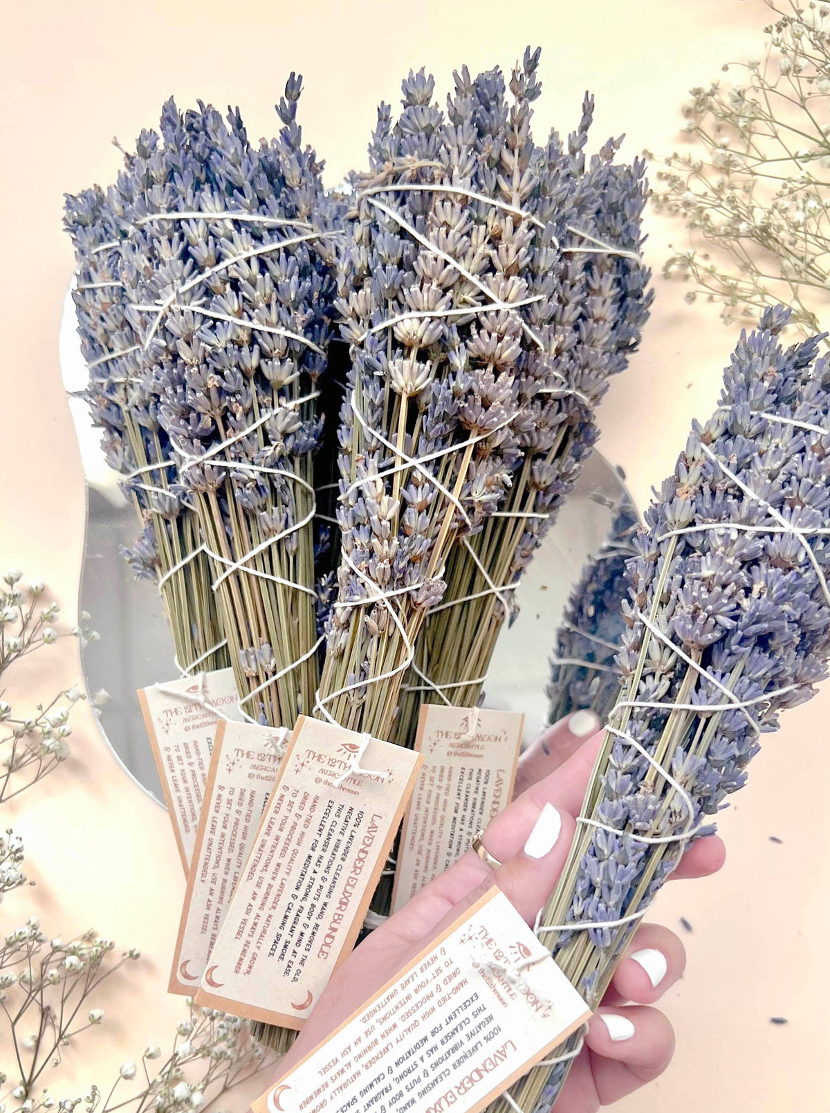 A hand holds several Lavender Elixir Smoke Cleansing Bundles by The 12th Moon, tied with white string and information cards. More bundles and small white flowers are scattered on a light background.