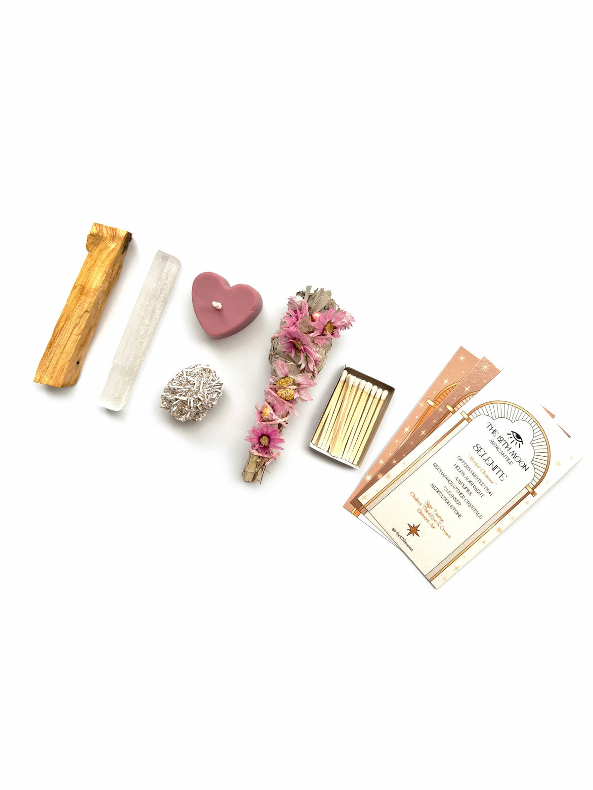 A flat lay of The 12th Moon’s Goddess Intentions Self-Care Ritual Kit, featuring a Palo Santo stick, selenite wand, heart-shaped candle, white crystal, Prairie Sage Bundle with pink flowers, matches, and incense—radiating Divine Feminine Energy.