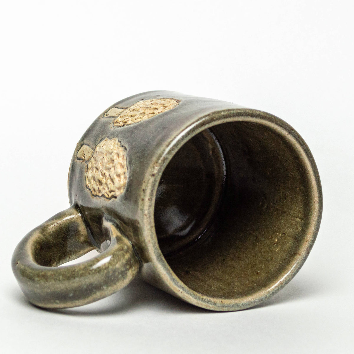 The Morel Mushroom Design Ceramic Mug by White Squirrel Clayworks is a 10oz handmade ceramic mug with a greenish-brown glaze and textured mushroom details, shown lying on its side against a plain white background.
