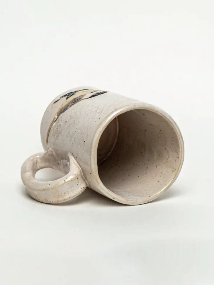 The Chickadee Design Handmade Ceramic Mug by White Squirrel Clayworks lies on its side on a white background, featuring a round hollow handle, speckled off-white glaze, and subtle black and tan design near the rim. 16oz capacity.