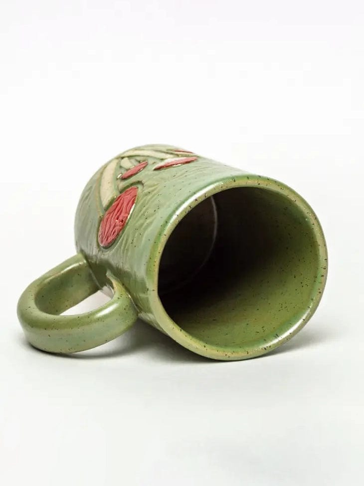 The Mushroom Design Handmade Ceramic Mug by White Squirrel Clayworks, a 16oz green ceramic mug with red accents and a textured surface, rests on its side with the handle visible and interior empty on a plain white background.
